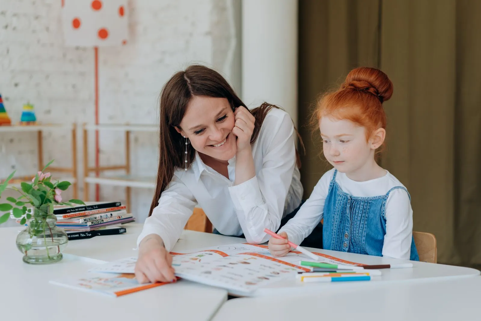 A smiling teacher assists a young girl with a colorful educational activity at a classroom table.
